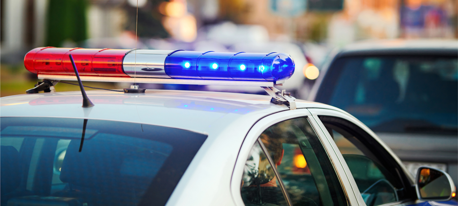 A close-up of a police car’s roof with its red and blue lights flashing. The background shows a blurred city street with other vehicles, suggesting the police car is responding to an incident or patrolling in an urban area. The image captures the sense of urgency and authority associated with law enforcement in action.