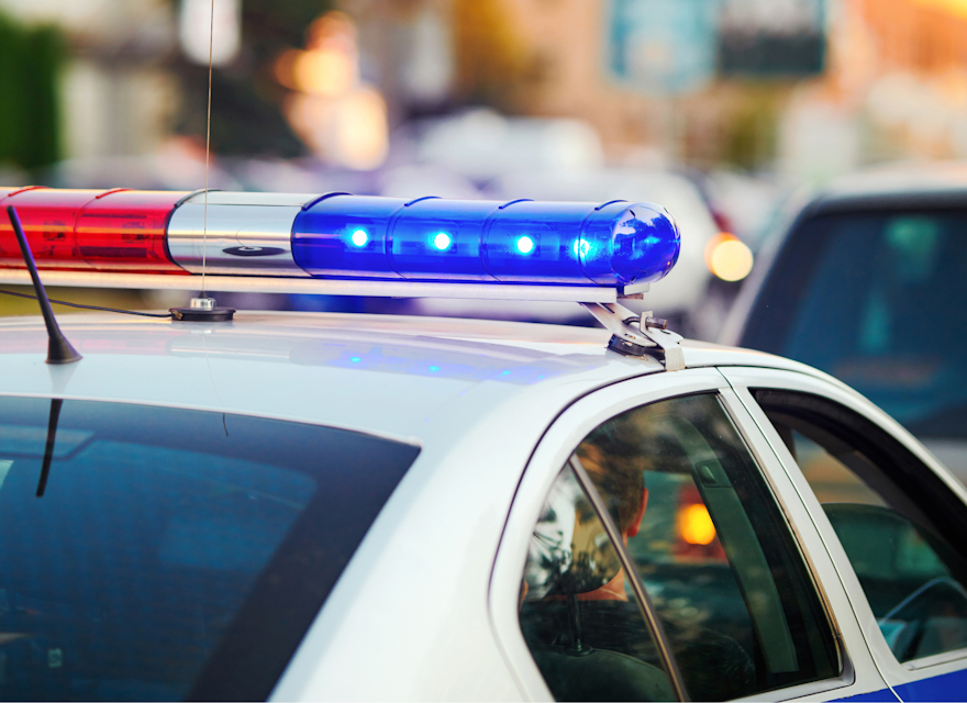 A close-up of a police car’s roof with its red and blue lights flashing. The background shows a blurred city street with other vehicles, suggesting the police car is responding to an incident or patrolling in an urban area. The image captures the sense of urgency and authority associated with law enforcement in action.