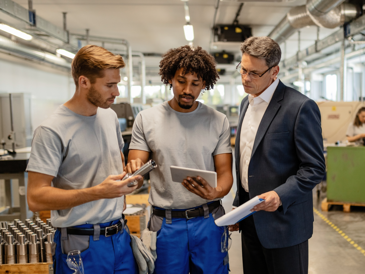 Three men discussing manufacturing equipment on a factory floor, with one holding a metal part, another using a tablet, and a third in a suit observing and holding a clipboard.