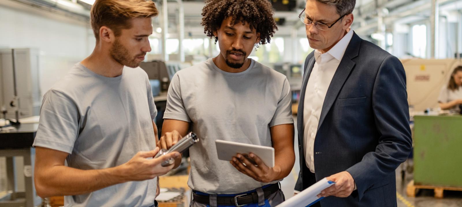 Three men discussing manufacturing equipment on a factory floor, with one holding a metal part, another using a tablet, and a third in a suit observing and holding a clipboard.