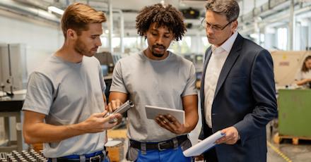 Three men discussing manufacturing equipment on a factory floor, with one holding a metal part, another using a tablet, and a third in a suit observing and holding a clipboard.