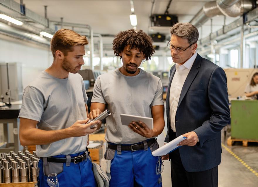 Three men discussing manufacturing equipment on a factory floor, with one holding a metal part, another using a tablet, and a third in a suit observing and holding a clipboard.