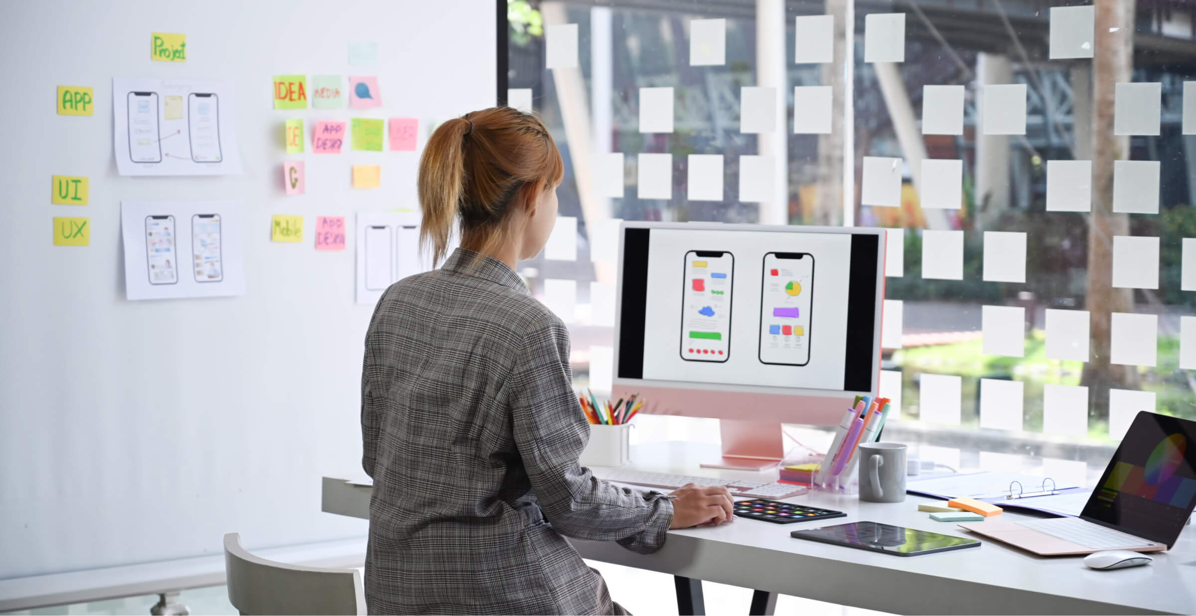 A designer working on a mobile app interface, sitting at a desk with a computer displaying app mockups. The workspace is organized with sticky notes on the wall, sketches, and design concepts. The bright and creative environment emphasizes the app development and design process.