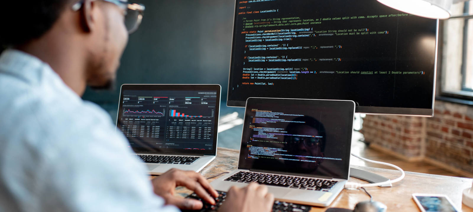 A software developer working at a desk with multiple screens, writing and analyzing code. The main monitor displays code in a text editor, while the laptop screens show graphs and additional coding windows. The workspace has a modern, focused atmosphere.
