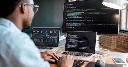 A software developer working at a desk with multiple screens, writing and analyzing code. The main monitor displays code in a text editor, while the laptop screens show graphs and additional coding windows. The workspace has a modern, focused atmosphere.