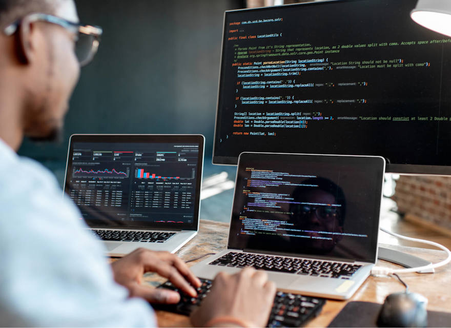 A software developer working at a desk with multiple screens, writing and analyzing code. The main monitor displays code in a text editor, while the laptop screens show graphs and additional coding windows. The workspace has a modern, focused atmosphere.