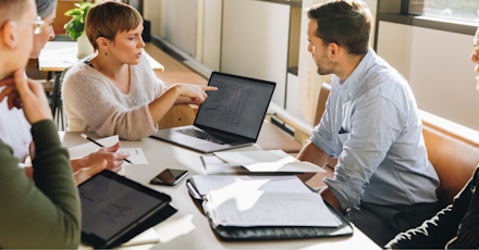 A group of five people is engaged in a discussion around a table with laptops