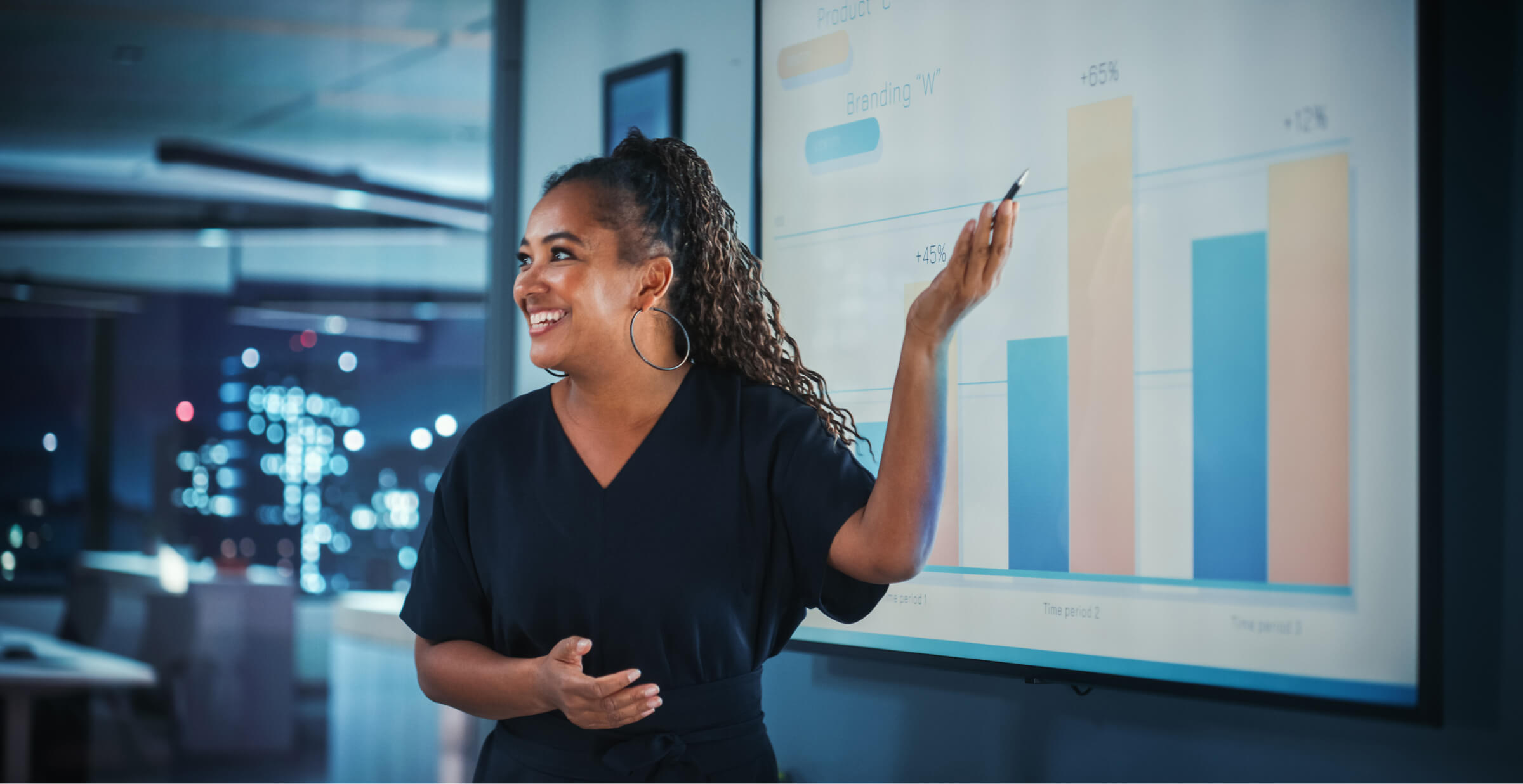 A woman stands in front of a projected presentation in a modern office, smiling and gesturing towards a bar chart with a pointer. She is wearing hoop earrings and a dark blouse. The background shows a cityscape through the window, illuminated with bright lights, suggesting it is evening. The chart displays percentages and data trends, indicating a professional business presentation.