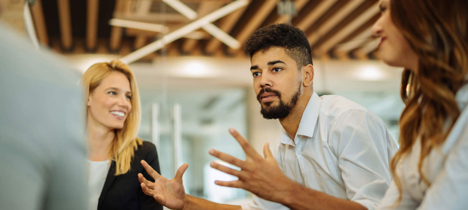 A group of colleagues in a modern office engaged in a discussion. A man with curly hair and a beard gestures with his hands while speaking, and two women, one with blonde hair and the other with brown, listen attentively. The background features a ceiling with wooden beams and contemporary lighting, creating a collaborative and dynamic atmosphere. The team appears focused on exchanging ideas, likely about AI