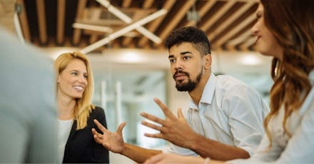 A group of colleagues in a modern office engaged in a discussion. A man with curly hair and a beard gestures with his hands while speaking, and two women, one with blonde hair and the other with brown, listen attentively. The background features a ceiling with wooden beams and contemporary lighting, creating a collaborative and dynamic atmosphere. The team appears focused on exchanging ideas, likely about AI