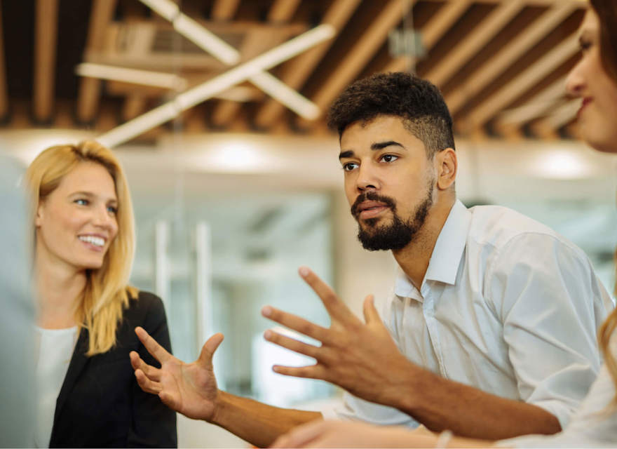 A group of colleagues in a modern office engaged in a discussion. A man with curly hair and a beard gestures with his hands while speaking, and two women, one with blonde hair and the other with brown, listen attentively. The background features a ceiling with wooden beams and contemporary lighting, creating a collaborative and dynamic atmosphere. The team appears focused on exchanging ideas, likely about AI