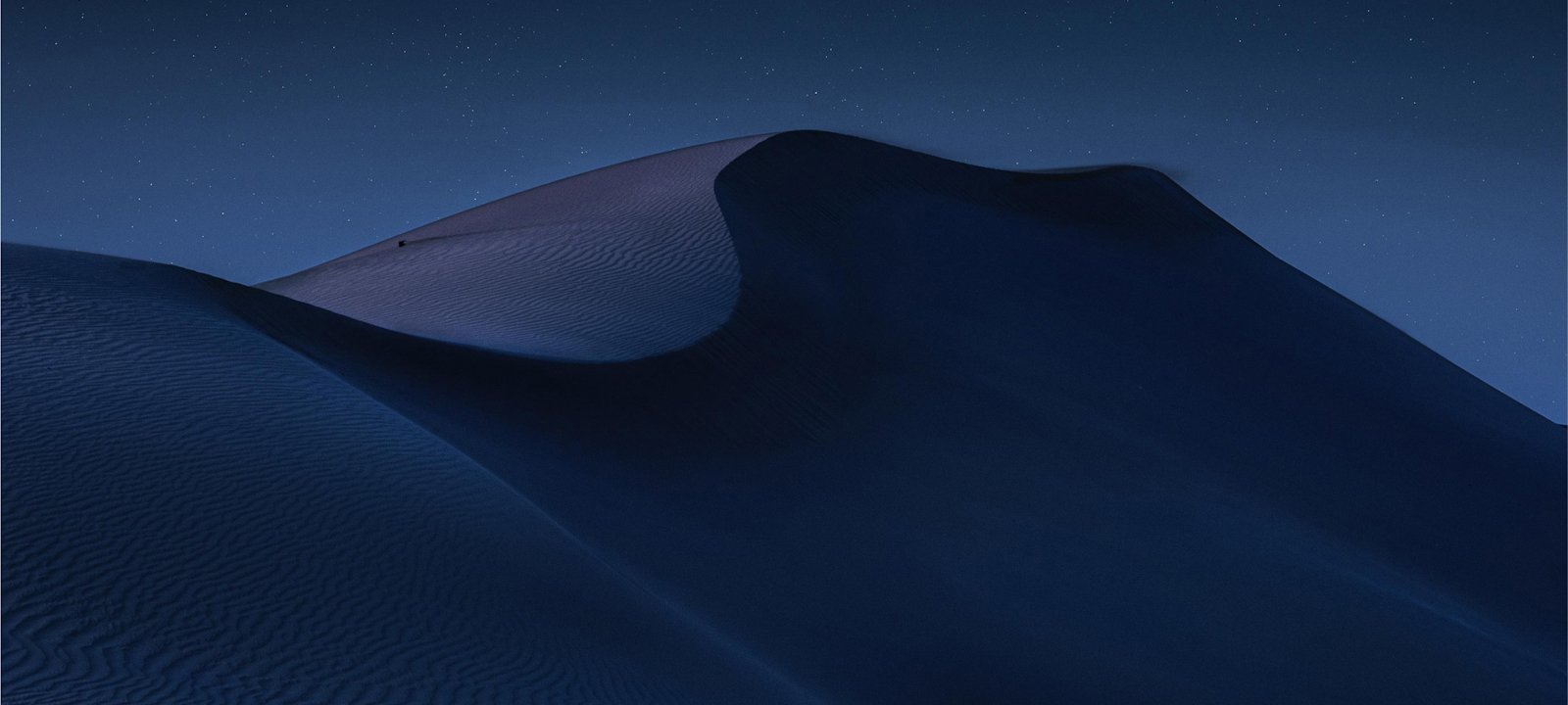 A serene nighttime landscape of desert dunes, softly illuminated under a clear, starry sky. The smooth curves and shadows of the sand create a dramatic, peaceful scene. The image captures the vastness and beauty of the desert under the calmness of night, evoking a sense of solitude and tranquility.