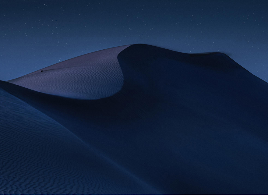 A serene nighttime landscape of desert dunes, softly illuminated under a clear, starry sky. The smooth curves and shadows of the sand create a dramatic, peaceful scene. The image captures the vastness and beauty of the desert under the calmness of night, evoking a sense of solitude and tranquility.