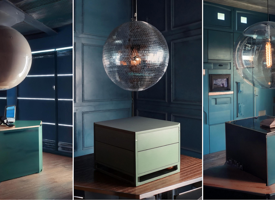 A modern office space featuring dark teal walls and minimalist green furniture, with large, spherical light fixtures hanging from the ceiling above desks and tables.