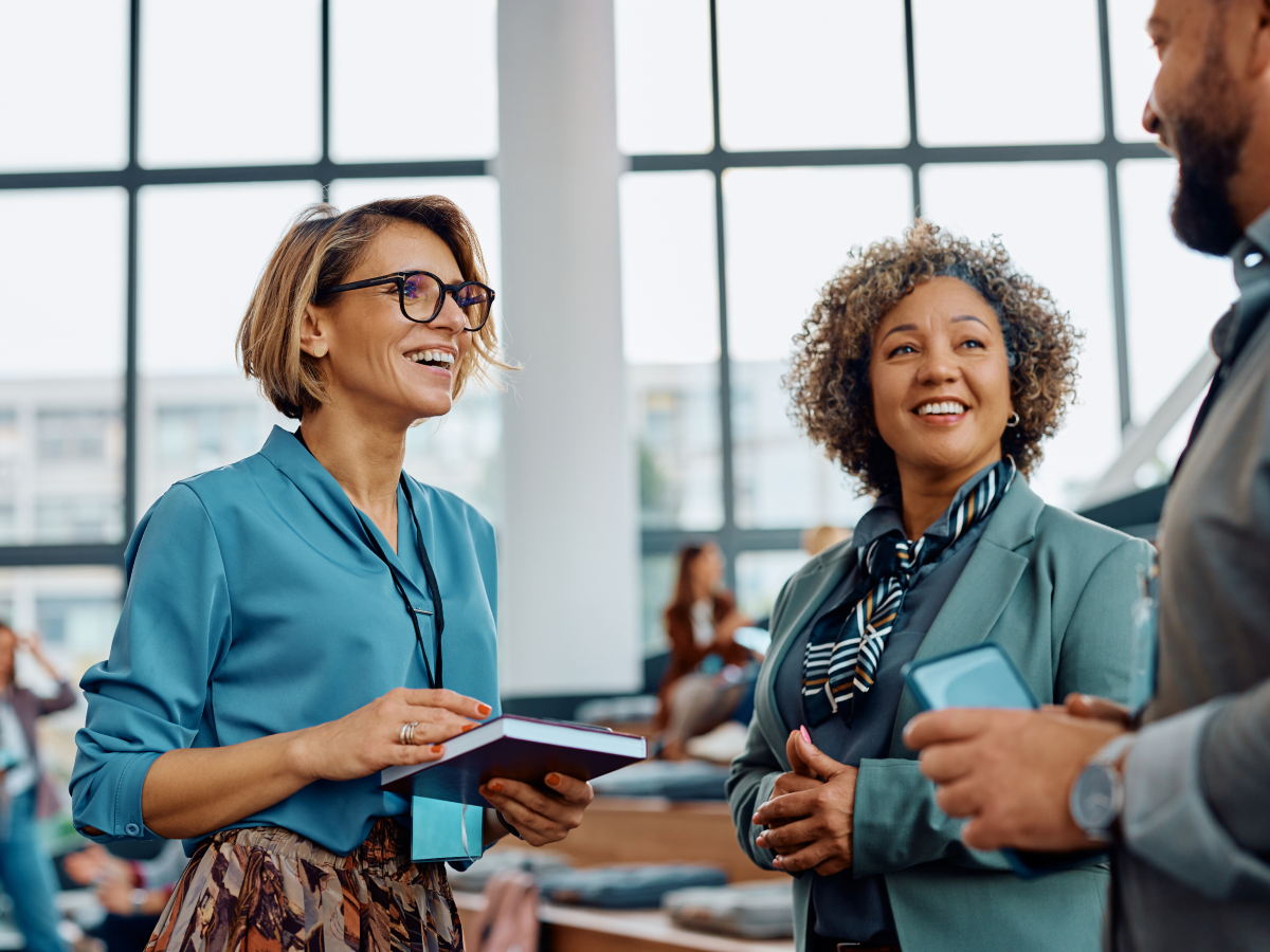 Two professional women smiling and conversing with a colleague in a bright, modern office setting with large windows in the background.