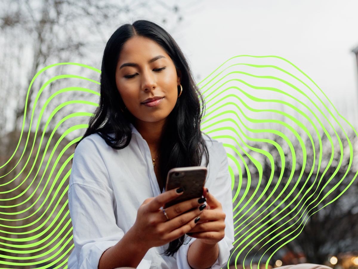 A woman sits outdoors, looking at her phone, dressed in a white shirt and beige pants, with sneakers
