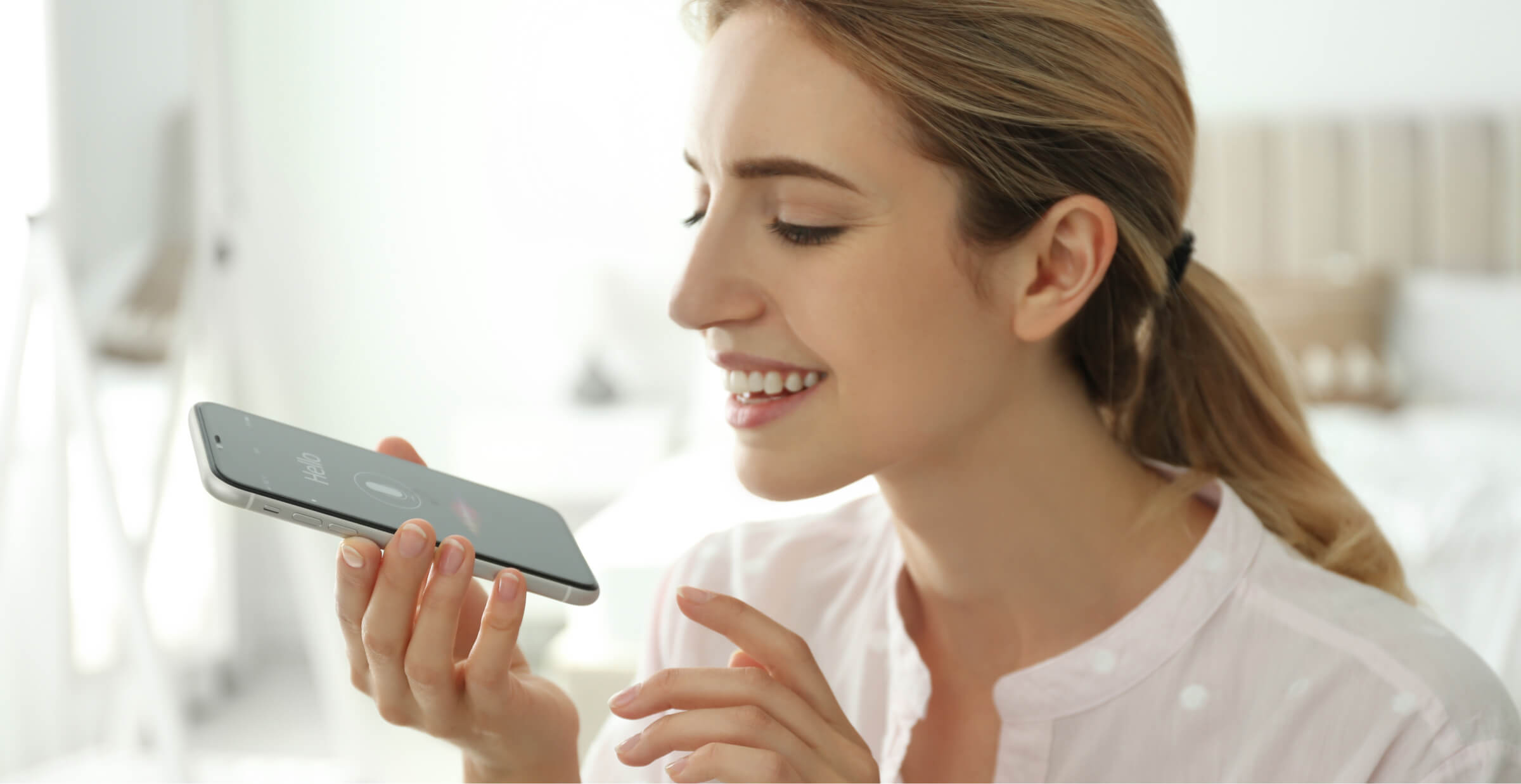 Woman smiling using voice command on smartphone in a bright room, illustrating modern AI interaction.