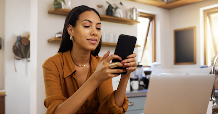 A woman is sitting in a cozy home office, smiling as she uses her smartphone. She appears relaxed and focused, with a laptop in front of her and a modern kitchen in the background. The setting suggests a comfortable and productive work-from-home environment, with natural light streaming through the windows. The image highlights the balance of professional and personal life in a home office setting.