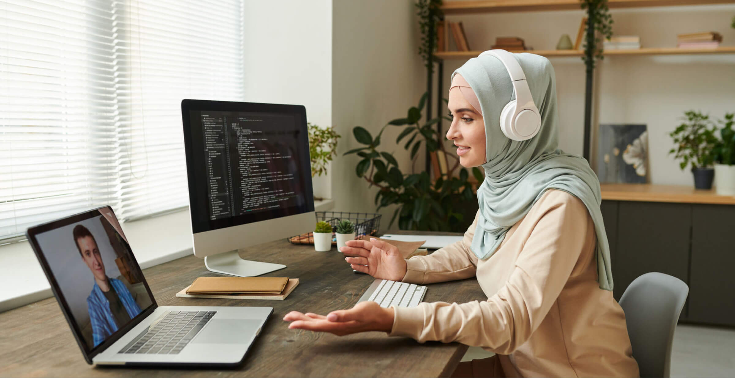 A woman wearing a hijab and headphones working from home, participating in a video call on her laptop while coding on a desktop computer. She is seated at a desk in a modern home office with plants and shelves in the background, engaged in a remote work setting.