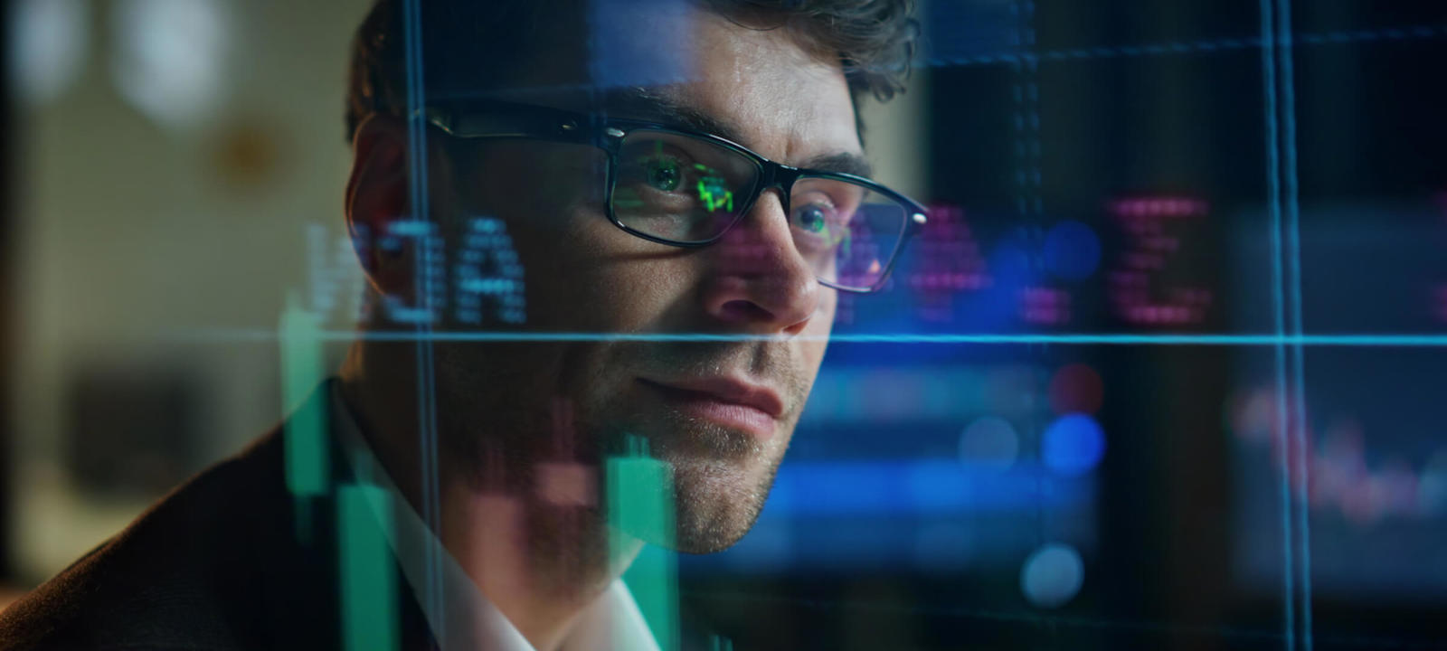 Man wearing glasses analyzing stock market data displayed on a digital screen with graphs and charts