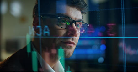 Man wearing glasses analyzing stock market data displayed on a digital screen with graphs and charts