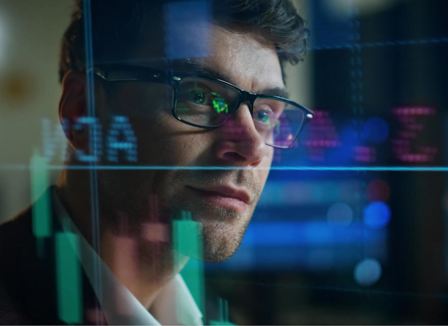 Man wearing glasses analyzing stock market data displayed on a digital screen with graphs and charts