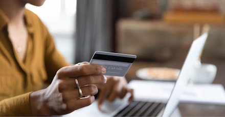 A close-up of a person holding a credit card while making an online purchase using a laptop. The person is typing with one hand and holding the card with the other, suggesting an online transaction or banking activity. The scene conveys the convenience and efficiency of digital financial services in a modern setting.