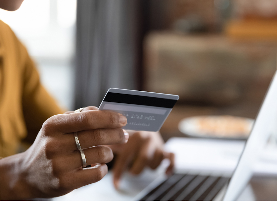 A close-up of a person holding a credit card while making an online purchase using a laptop. The person is typing with one hand and holding the card with the other, suggesting an online transaction or banking activity. The scene conveys the convenience and efficiency of digital financial services in a modern setting.
