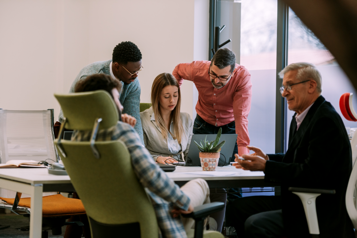A group of five people, three men and two women, are gathered around a laptop in an office setting, engaged in a discussion. One man and one woman are seated, while the others stand.