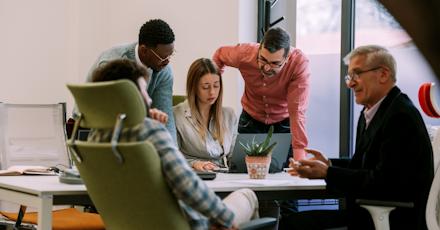 A group of five people, three men and two women, are gathered around a laptop in an office setting, engaged in a discussion. One man and one woman are seated, while the others stand.