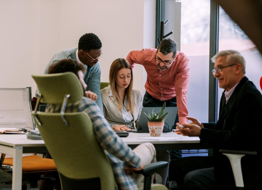 A group of five people, three men and two women, are gathered around a laptop in an office setting, engaged in a discussion. One man and one woman are seated, while the others stand.
