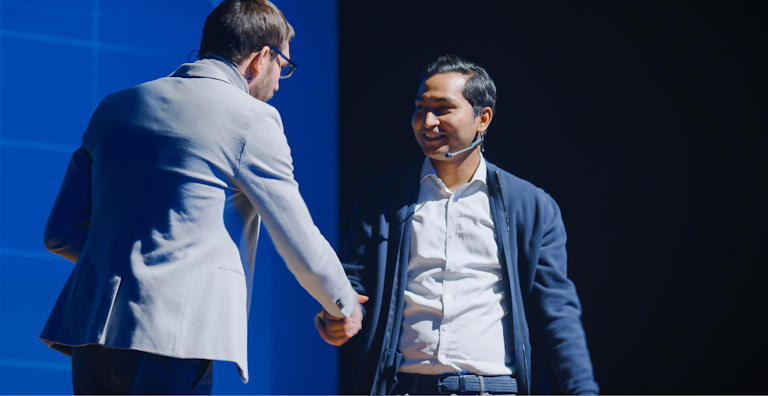 Two me, one in a gray suit and the other in a dark jacket, shaking hands on stage during a conference or event.