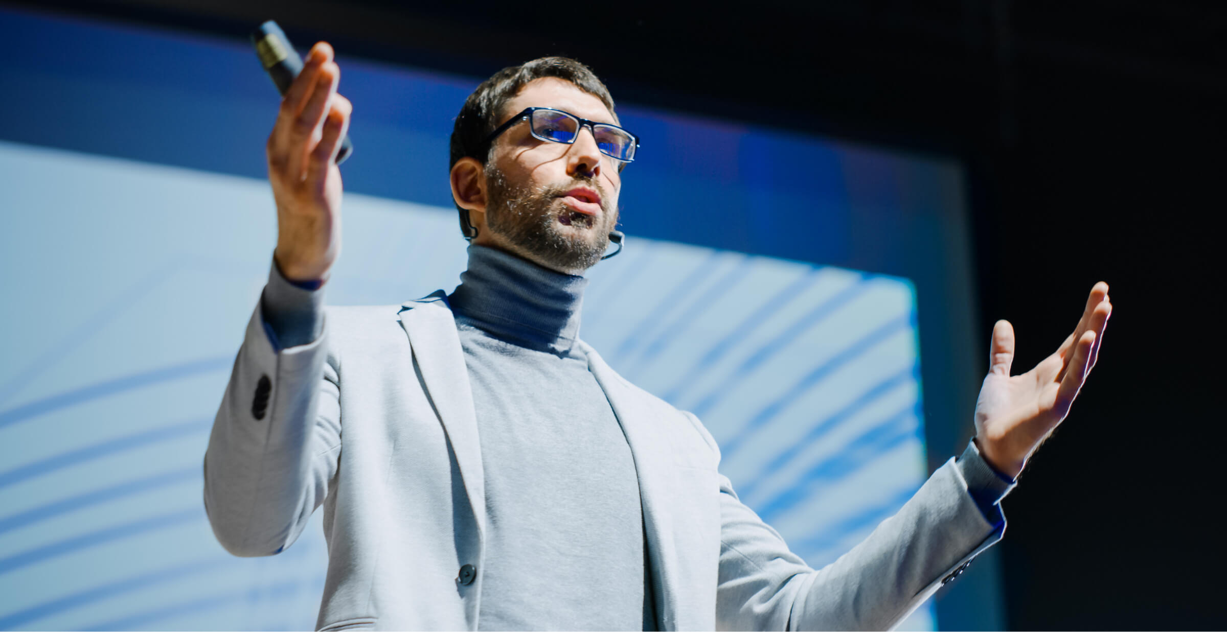 A man delivering a presentation on stage with a microphone, wearing a suit and glasses.