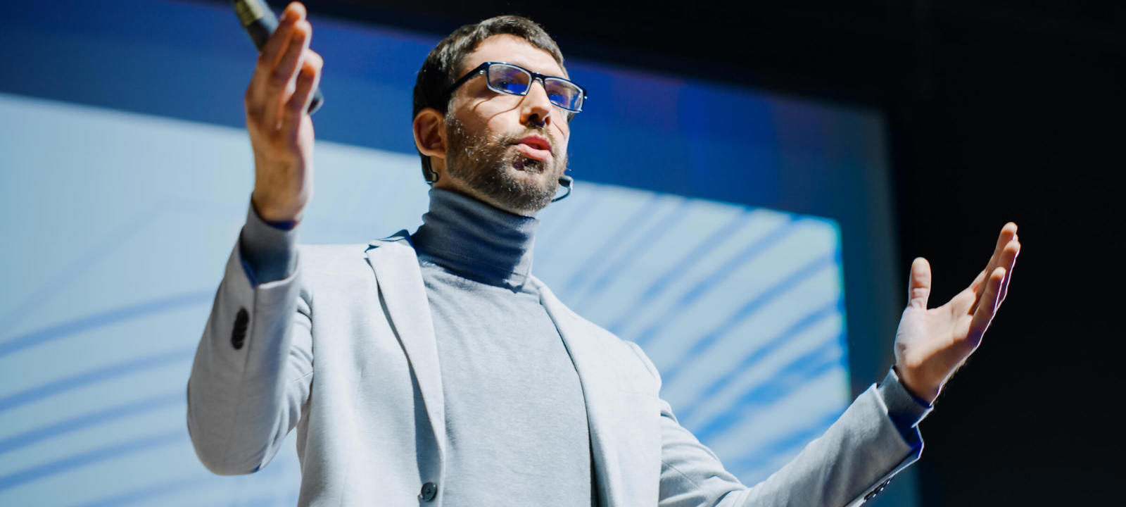 A man delivering a presentation on stage with a microphone, wearing a suit and glasses.