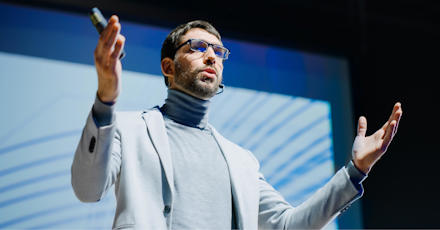 A man delivering a presentation on stage with a microphone, wearing a suit and glasses.