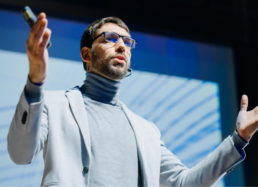 A man delivering a presentation on stage with a microphone, wearing a suit and glasses.