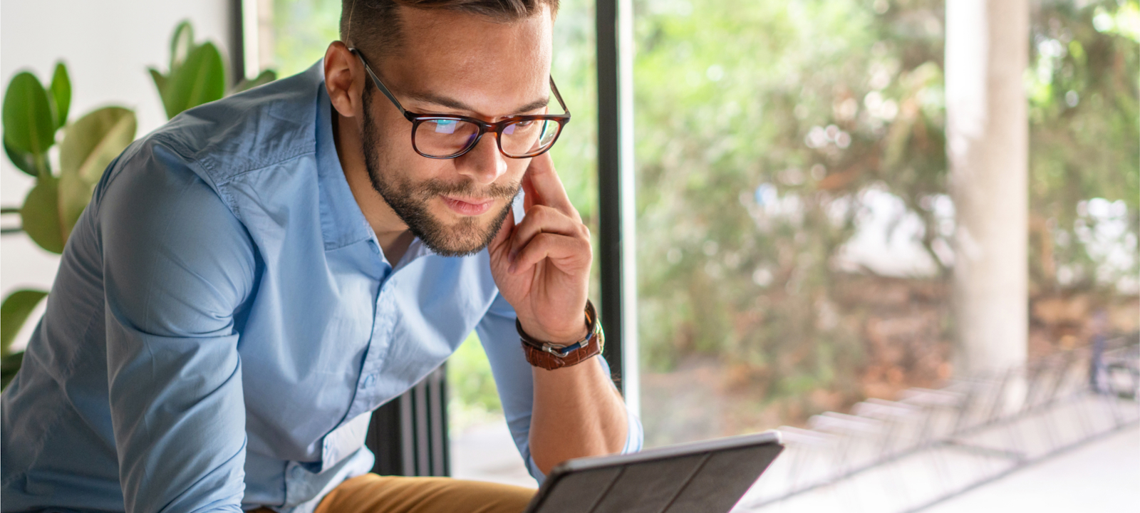 A young man wearing glasses and a light blue shirt is sitting by a window, focused on working on a tablet. He appears engaged, possibly in a video call or reviewing information. The background shows greenery and natural light, creating a calm and productive workspace. The image conveys a sense of modern, remote work in a comfortable and professional environment.