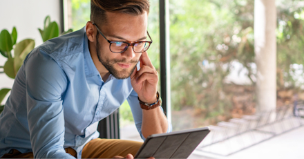 A young man wearing glasses and a light blue shirt is sitting by a window, focused on working on a tablet. He appears engaged, possibly in a video call or reviewing information. The background shows greenery and natural light, creating a calm and productive workspace. The image conveys a sense of modern, remote work in a comfortable and professional environment.