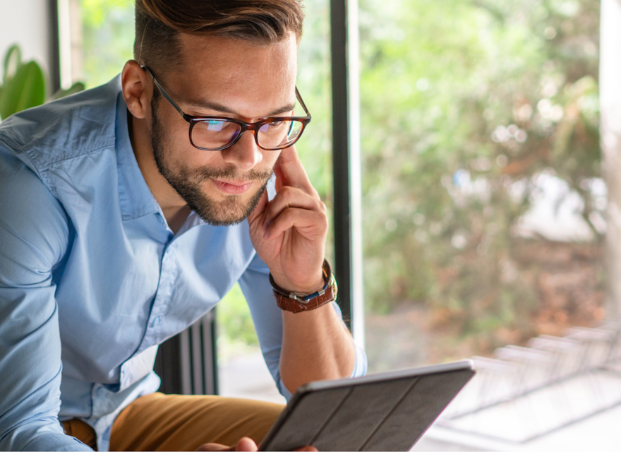 A young man wearing glasses and a light blue shirt is sitting by a window, focused on working on a tablet. He appears engaged, possibly in a video call or reviewing information. The background shows greenery and natural light, creating a calm and productive workspace. The image conveys a sense of modern, remote work in a comfortable and professional environment.