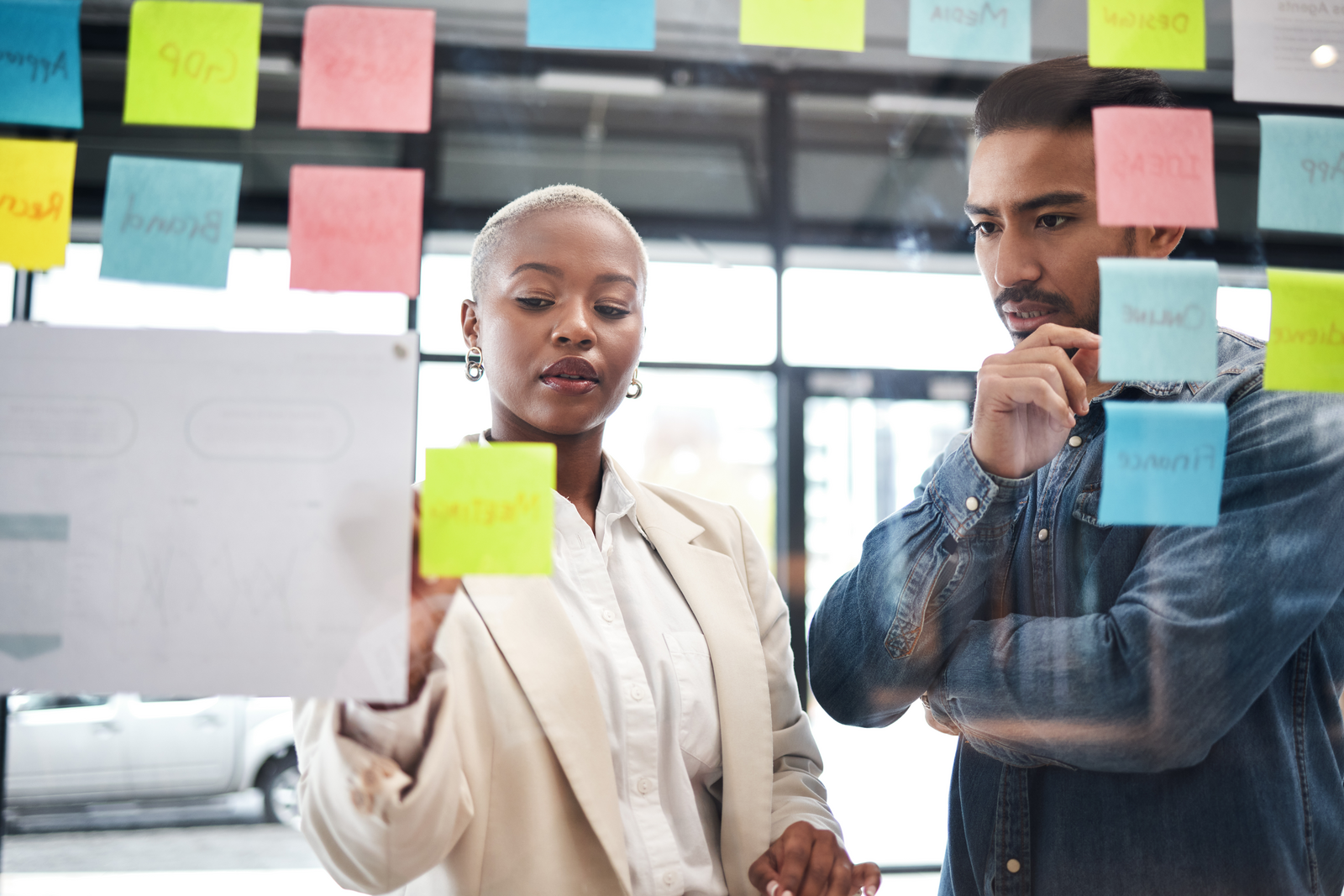 Two professionals are engaged in a collaborative brainstorming session, using a glass wall covered with colorful sticky notes. The woman, with short blonde hair and wearing a beige blazer, is placing a yellow sticky note on the wall, while the man next to her, dressed in a denim shirt, observes thoughtfully with his hand on his chin. 