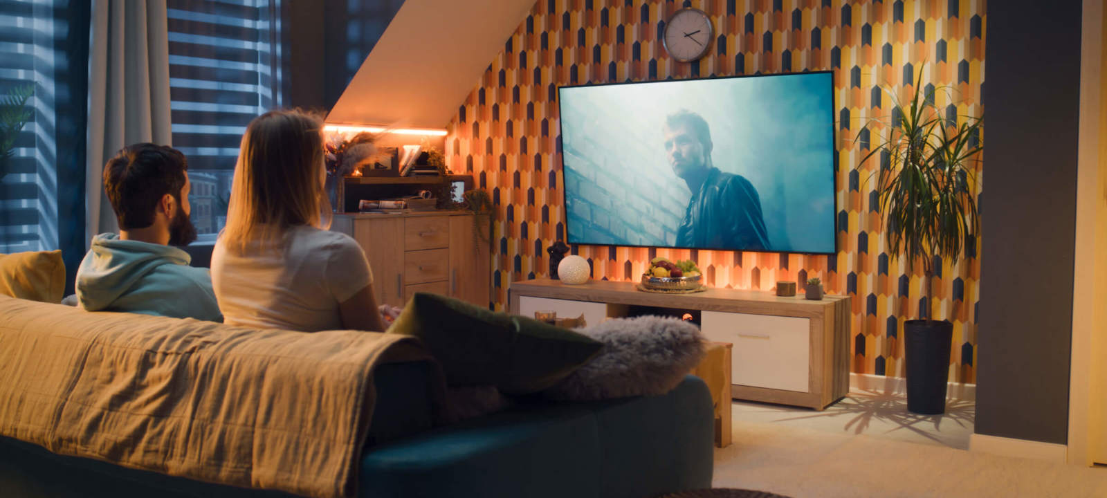 Couple sitting on a sofa in a modern living room, watching a movie on a large TV screen