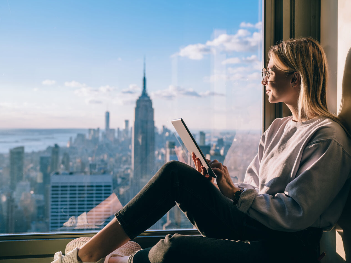 A woman with glasses sits by a large window, holding a tablet, while gazing out at the new york city skyline with the empire state building in view on a sunny day