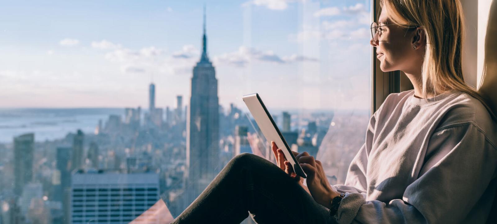 A woman with glasses sits by a large window, holding a tablet, while gazing out at the new york city skyline with the empire state building in view on a sunny day
