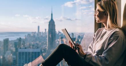 A woman with glasses sits by a large window, holding a tablet, while gazing out at the new york city skyline with the empire state building in view on a sunny day
