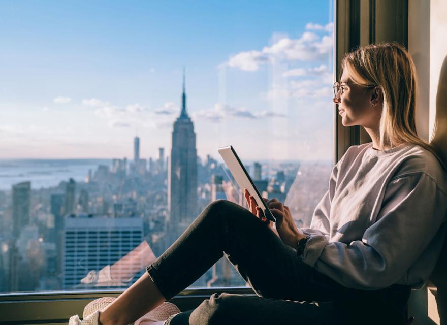 A woman with glasses sits by a large window, holding a tablet, while gazing out at the new york city skyline with the empire state building in view on a sunny day