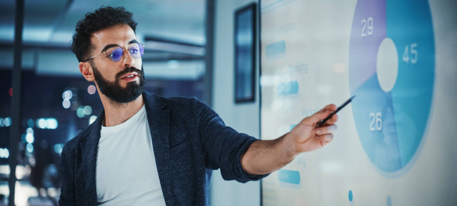 A man with glasses presents data on a screen, pointing to a pie chart during a business meeting.