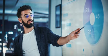 A man with glasses presents data on a screen, pointing to a pie chart during a business meeting.