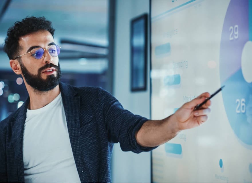 A man with glasses presents data on a screen, pointing to a pie chart during a business meeting.