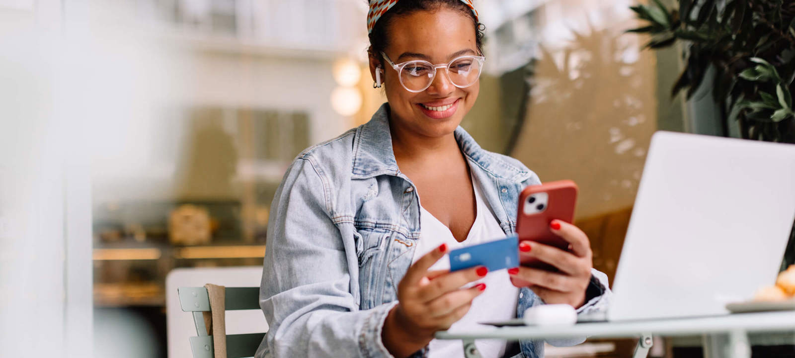A young woman sitting at a cafe table, smiling as she uses her smartphone and holds a credit card, with a laptop open in front of her. She is wearing a denim jacket, glasses, and a colorful headband, suggesting she is making an online purchase or transaction in a relaxed setting.