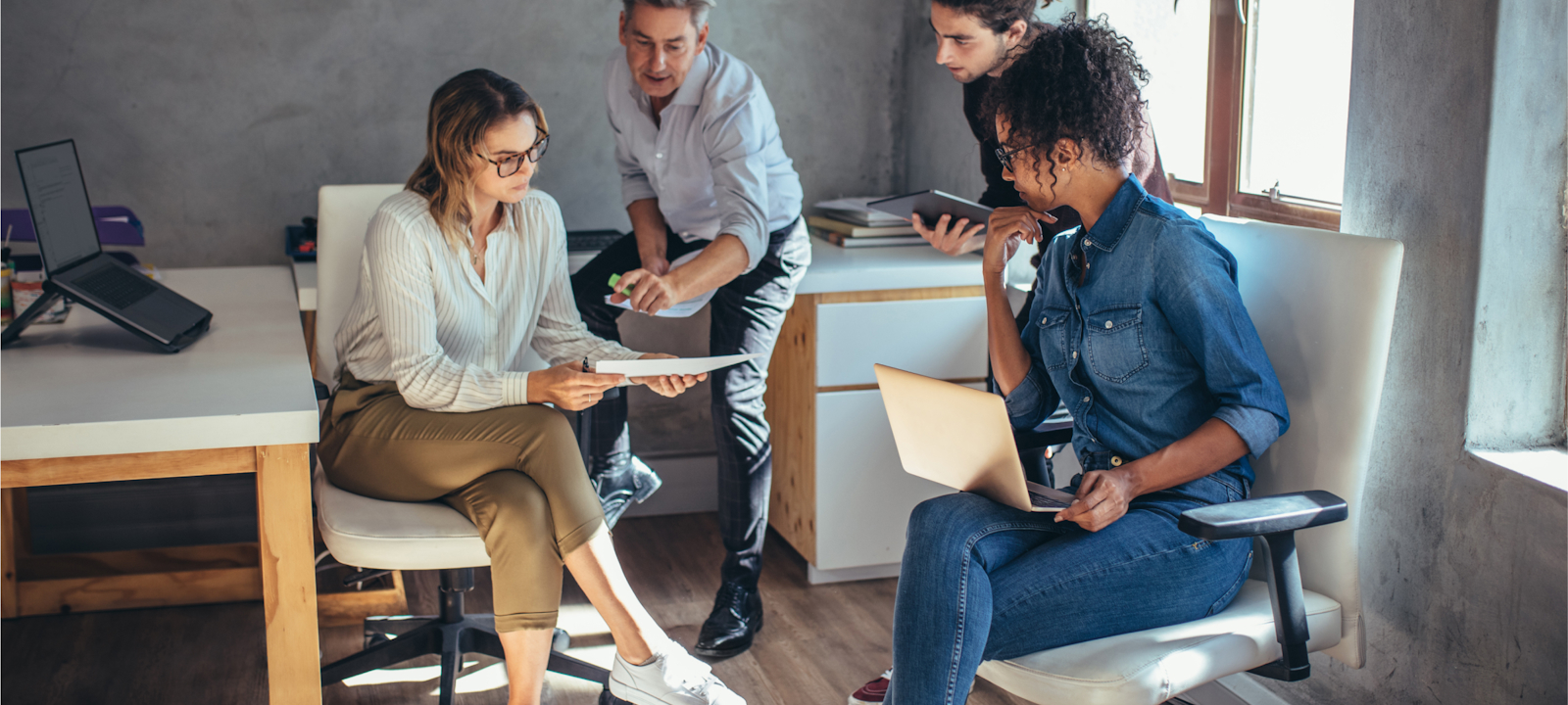 A group of four colleagues is engaged in a collaborative discussion in a modern office setting. One woman holds a document while another reviews it, with two other team members contributing to the conversation. Laptops and tablets are present, indicating a productive work session. The office environment is casual and bright, with natural light streaming through the windows, reflecting a focused and dynamic team effort.